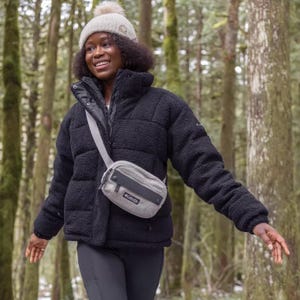 A woman wearing a black puffer jacket, beige beanie, and sporting a grey crossbody bag stands in a forest.
