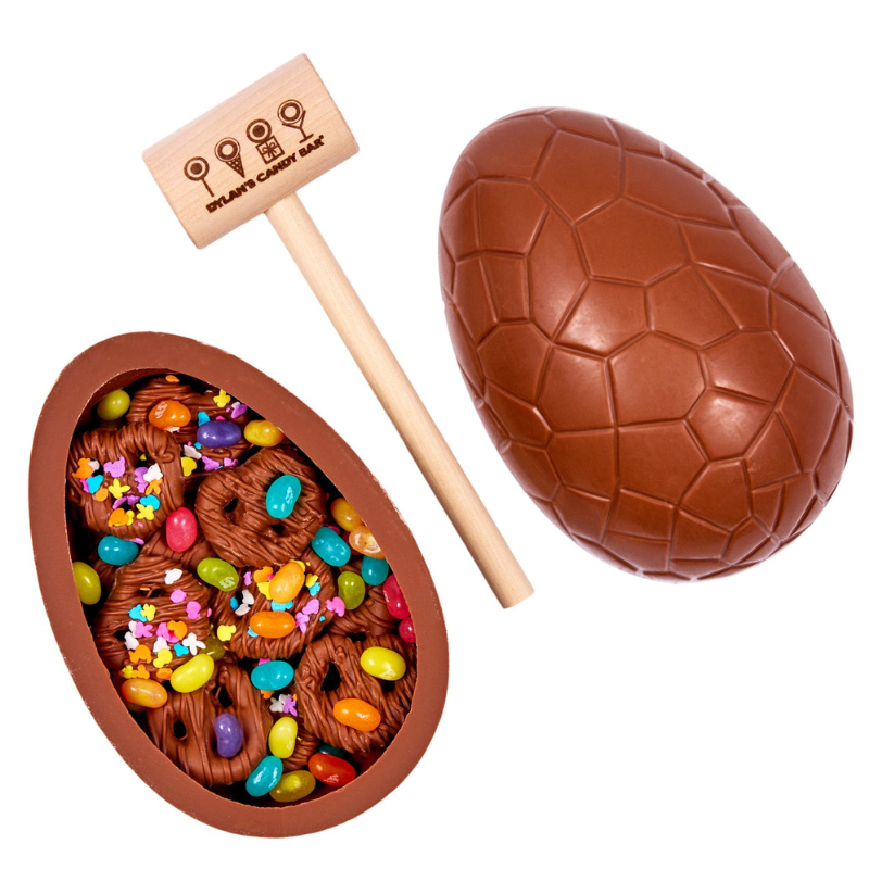 A breakable chocolate egg filled with colorful candies and chocolate-covered pretzels, accompanied by a small wooden mallet with the Dylan's Candy Bar logo.