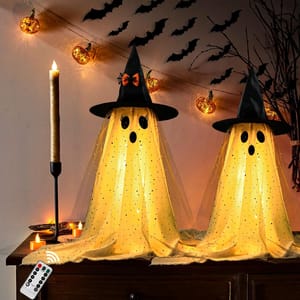 Two lighted ghost decorations wearing witch hats are set on a table, surrounded by Halloween-themed decor like bat cutouts, pumpkin string lights, a candle, and a remote control.