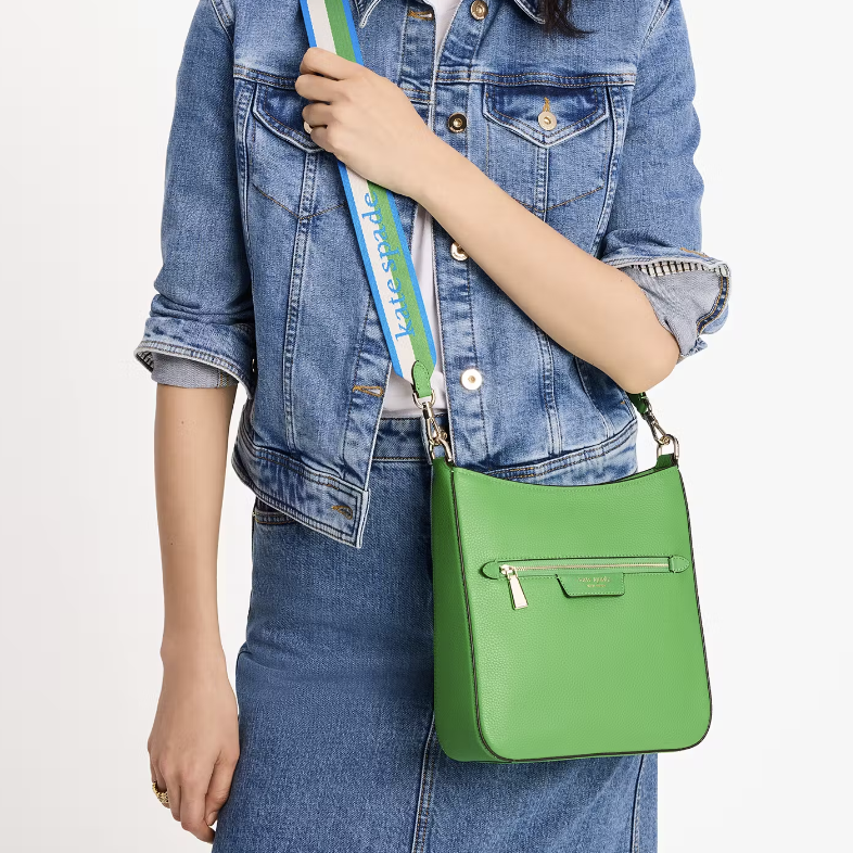 A person wearing a denim jacket and jeans holding a green shoulder bag with a blue strap featuring a logo.