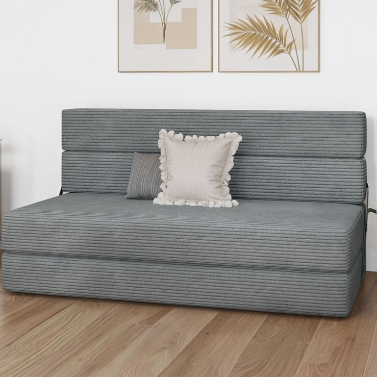A gray, foldable futon sofa bed with striped fabric is shown, featuring two decorative pillows. It is set against a white wall with two framed botanical prints.