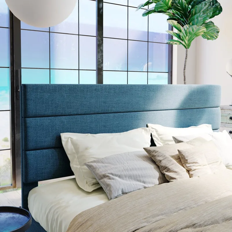 A bedroom scene with a blue upholstered headboard, beige and white pillows, and a beige comforter, positioned in front of large windows showcasing a view of the ocean. A tall green plant is on the right side.