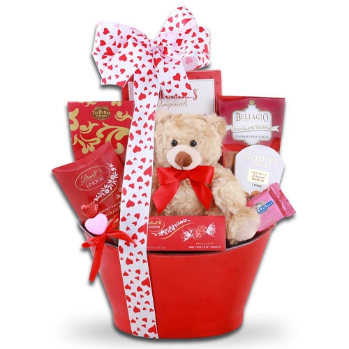 A red gift basket containing a teddy bear, assorted chocolates, and hot cocoa mixes, wrapped with a white and red heart-patterned ribbon. Brands include Lindt, Godiva, and Bellagio.
