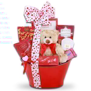 A red gift basket containing a teddy bear, assorted chocolates, and hot cocoa mixes, wrapped with a white and red heart-patterned ribbon. Brands include Lindt, Godiva, and Bellagio.