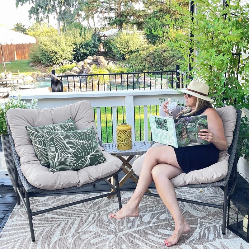 A woman relaxes on a patio chair with cushions, beside a table with a decorative yellow lantern, holding a glass and a magazine.