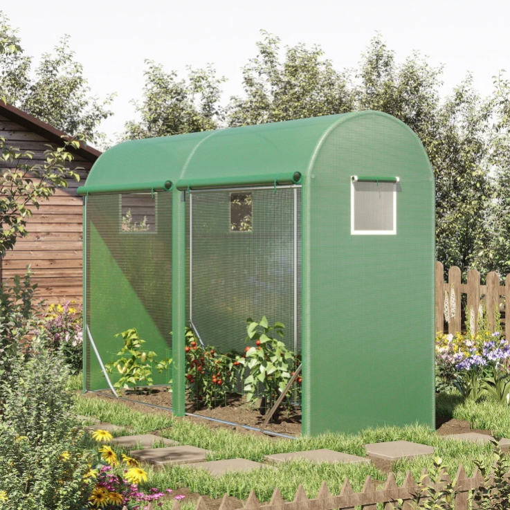 A small green greenhouse with arched roof and mesh windows is situated next to a wooden shed in a garden with various plants and flowers.