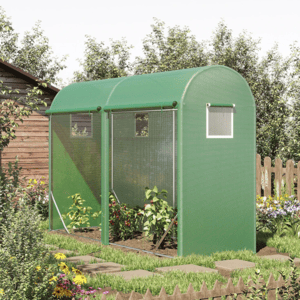 A small green greenhouse with arched roof and mesh windows is situated next to a wooden shed in a garden with various plants and flowers.