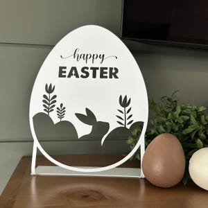 White Easter egg-shaped tabletop sign with \“Happy Easter\“ text, featuring a silhouette of a bunny and plants. It sits on a wooden surface next to brown and white egg-shaped decorations and greenery.