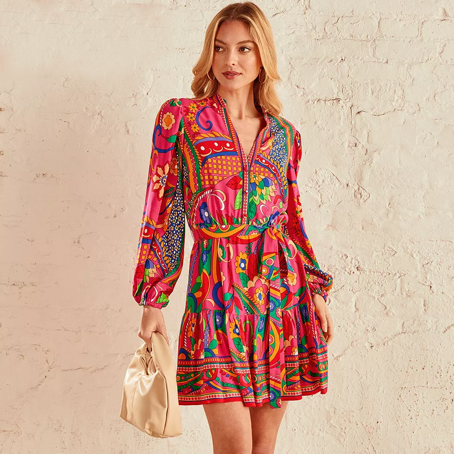 A woman is wearing a vibrant, multicolored dress with floral and geometric patterns, complemented by a pale gold clutch bag.