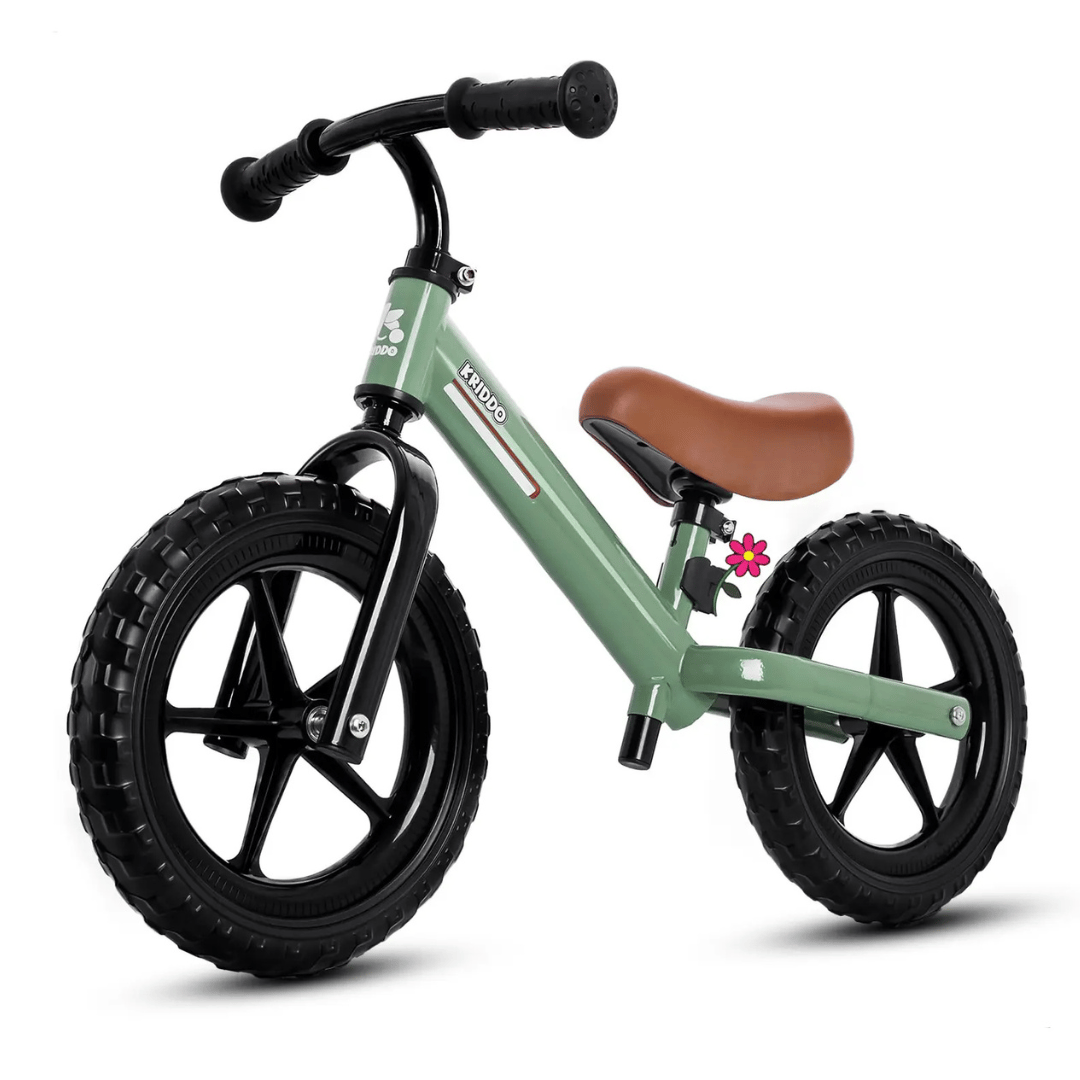 A green balance bike with black wheels and a brown seat, featuring a small pink flower detail near the seat post.