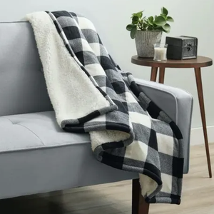 A black and white checkered throw blanket with a soft white interior is draped over a light gray sofa, next to a round wooden side table holding a plant, candle, and decorative box.