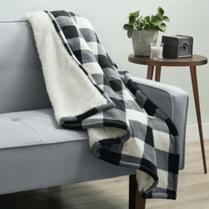 A black and white checkered throw blanket with a soft white interior is draped over a light gray sofa, next to a round wooden side table holding a plant, candle, and decorative box.