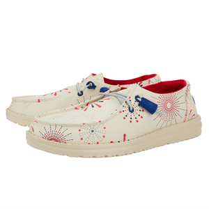Hey Dude Wendy Fireworks Loafers feature a beige canvas with red and blue fireworks patterns, red interior, and white soles. They are designed with light gray laces and blue lace holders for a casual, festive look.