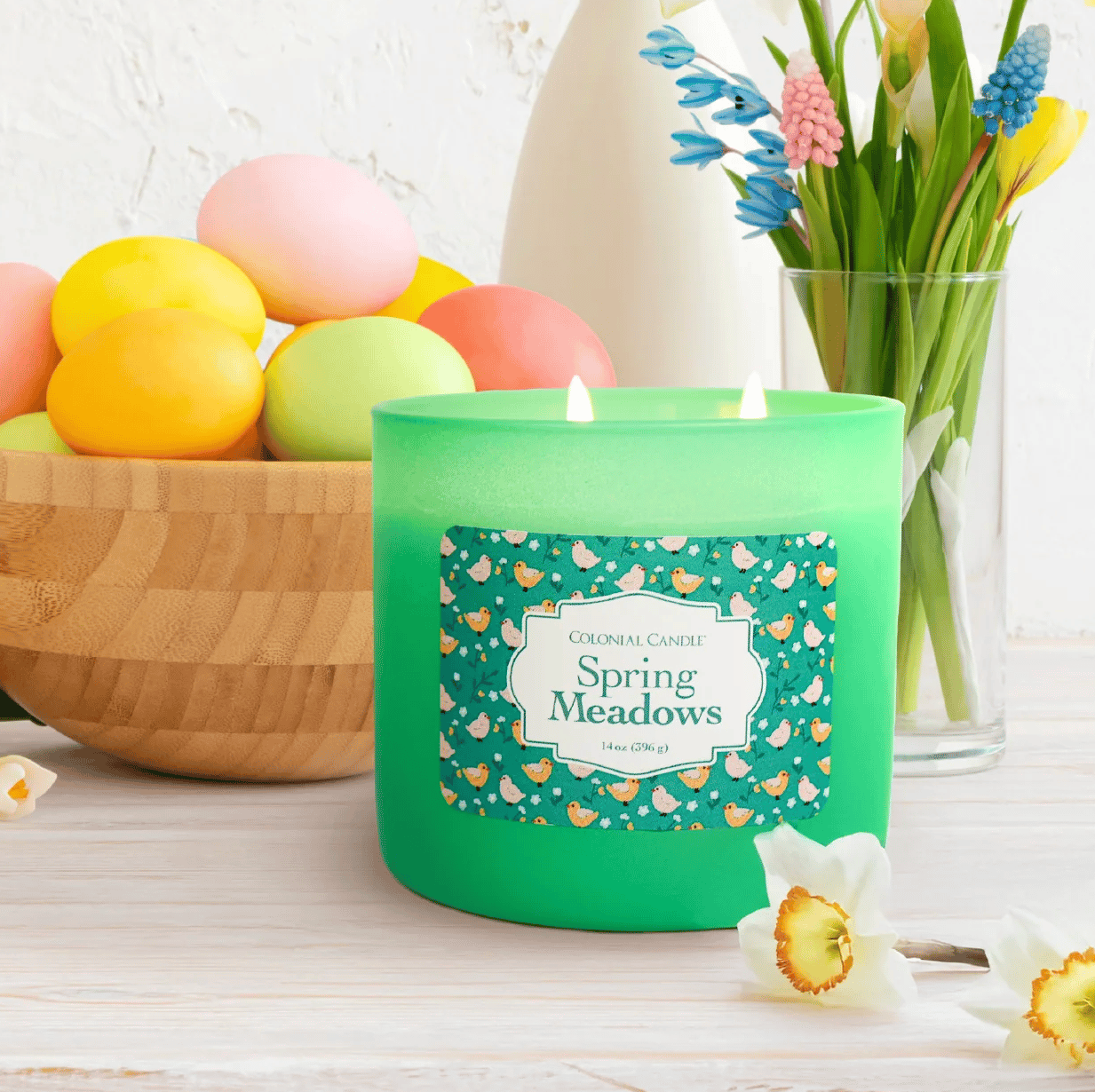 A green \“Spring Meadows\“ scented candle by Colonial Candle is lit and placed on a table next to a bowl of colorful Easter eggs and a vase of flowers.