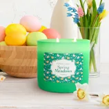 A green \“Spring Meadows\“ scented candle by Colonial Candle is lit and placed on a table next to a bowl of colorful Easter eggs and a vase of flowers.