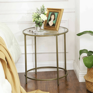 A round glass-top side table with a gold metal frame, featuring a vase with flowers and a framed portrait. It's positioned beside a bed with a light blanket draped over it, and a potted plant is nearby.