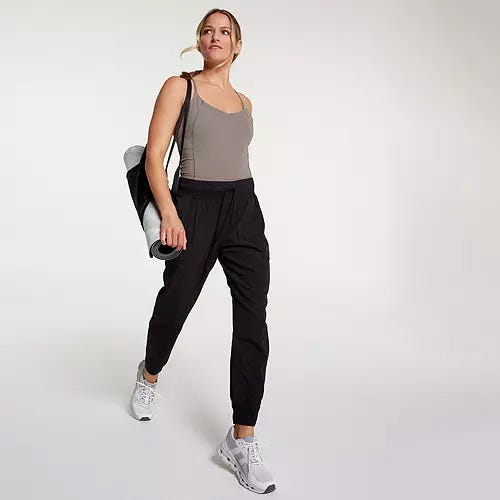 A woman wearing casual jogger pants, a tank top, sneakers, and carrying a yoga mat over her shoulder.