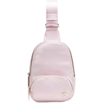 A small, light pink sling backpack with a zippered main compartment and a smaller front pocket, featuring a subtle logo emblem.
