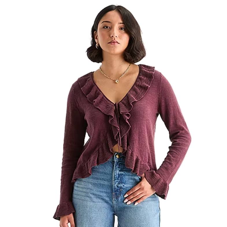 A woman is wearing a maroon long-sleeve top with ruffled detailing along the neckline and hem, paired with high-waisted light blue jeans.
