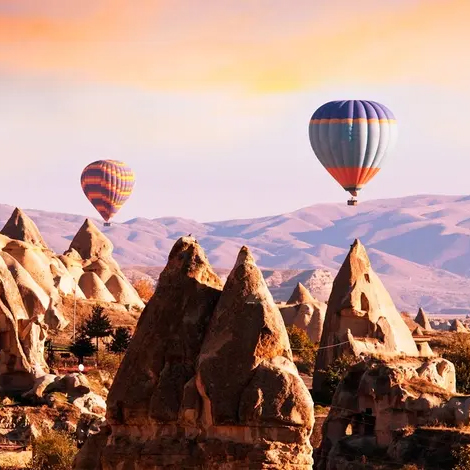 Hot air balloons float over unique rock formations and a scenic landscape with mountains in the background, during sunset.