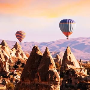 Hot air balloons float over unique rock formations and a scenic landscape with mountains in the background, during sunset.