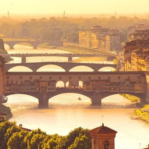 A scenic view of the Ponte Vecchio and the Arno River in Florence, Italy, with several bridges and historic buildings bathed in a golden sunset glow.