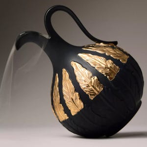 A black ceramic vase with an elegant curved handle, adorned with gold leaf-like patterns, featuring a side spout.