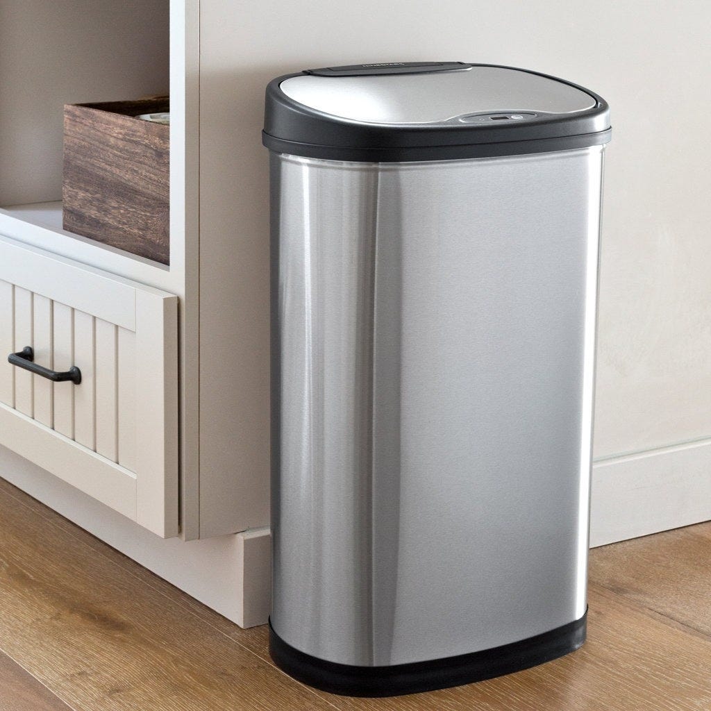 A stainless steel, rectangular, sensor-operated trash can with a black lid sits on a wooden floor next to a light-colored cabinet.