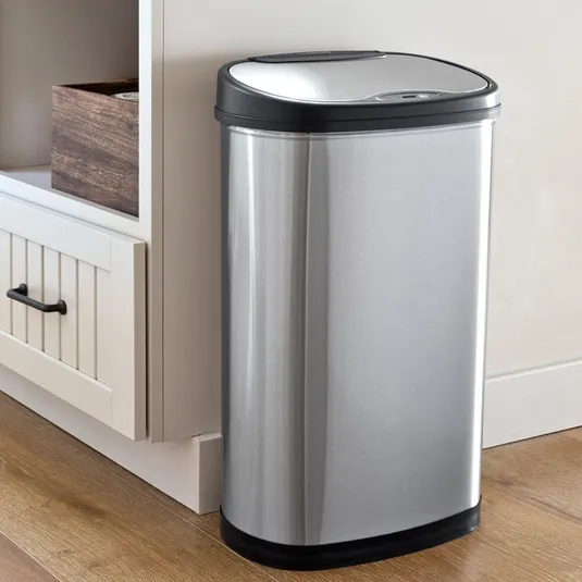 A stainless steel, rectangular, sensor-operated trash can with a black lid sits on a wooden floor next to a light-colored cabinet.