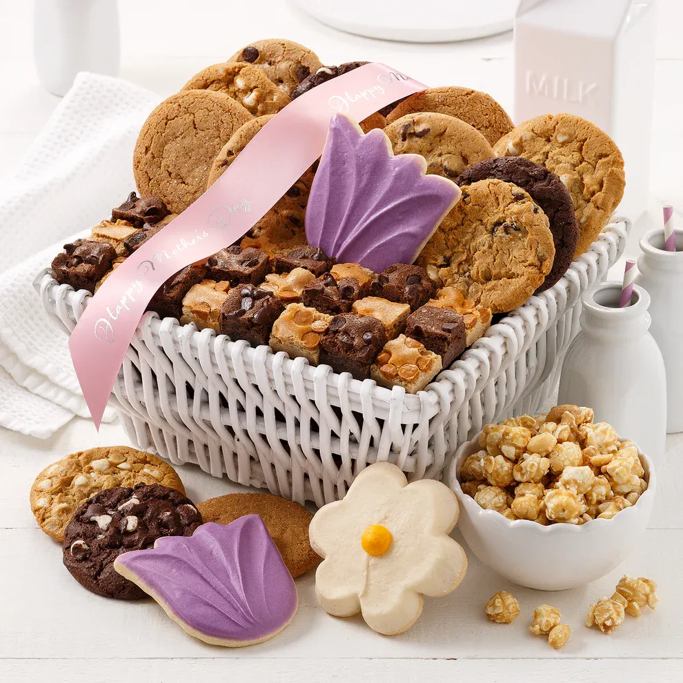 A white wicker basket filled with a variety of cookies, including chocolate chip, sugar cookies with purple and white icing, and brownies, accompanied by caramel popcorn. A pink ribbon reading \“Happy Mother's Day\“ is draped over the basket.