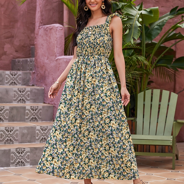 A woman wears a sleeveless, ankle-length floral dress with yellow and green patterns, featuring a smocked bodice. She stands next to pink walls and tropical plants on tiled steps.