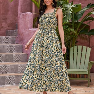 A woman wears a sleeveless, ankle-length floral dress with yellow and green patterns, featuring a smocked bodice. She stands next to pink walls and tropical plants on tiled steps.