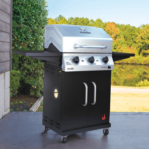 A stainless steel Char-Broil gas grill with three control knobs, side shelves, and a cabinet base with two doors, positioned outdoors near a lake and greenery.
