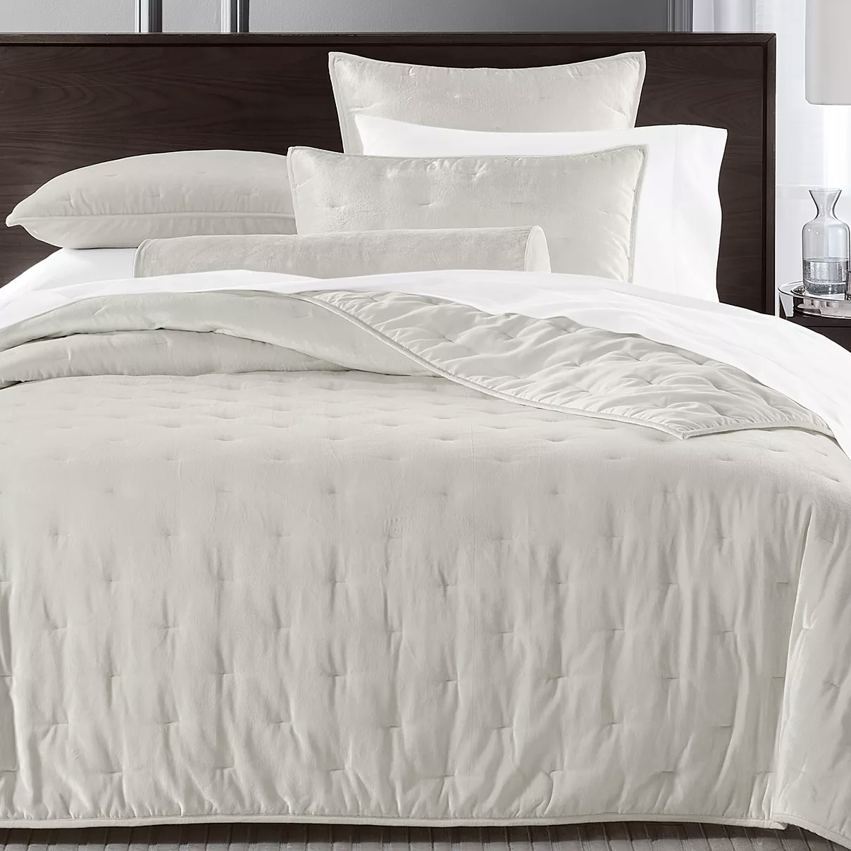 A neatly made bed showcases a set of creamy white linens, including a comforter, pillows, and a bolster cushion, complemented by a dark wooden headboard. A clear glass and a small decor item rest on the bedside table.