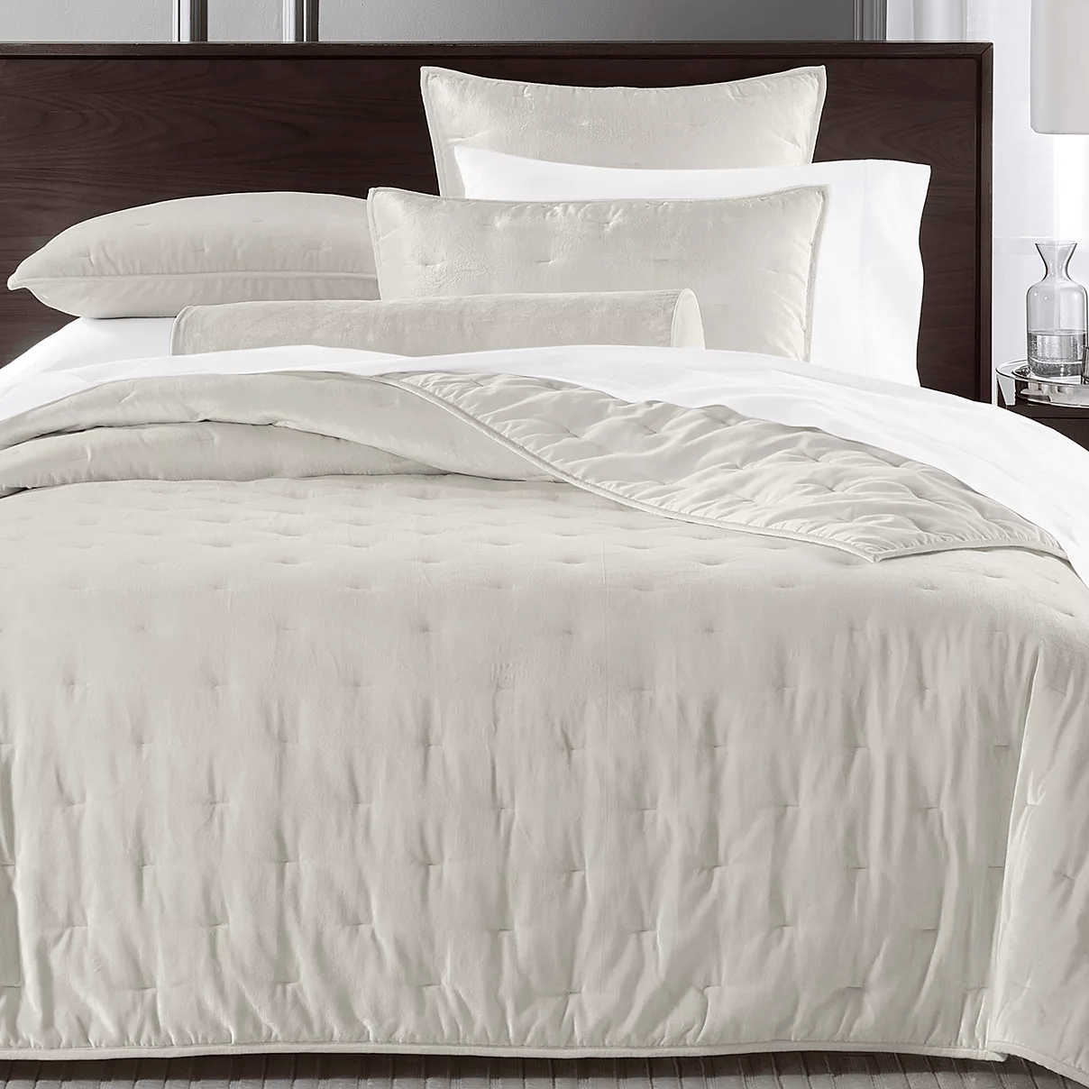 A neatly made bed showcases a set of creamy white linens, including a comforter, pillows, and a bolster cushion, complemented by a dark wooden headboard. A clear glass and a small decor item rest on the bedside table.
