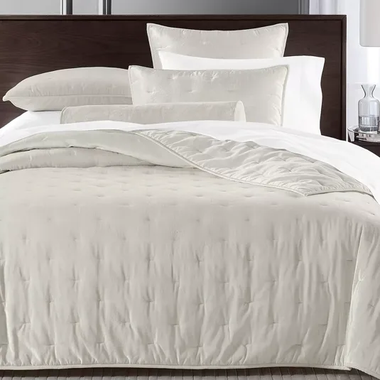 A neatly made bed showcases a set of creamy white linens, including a comforter, pillows, and a bolster cushion, complemented by a dark wooden headboard. A clear glass and a small decor item rest on the bedside table.