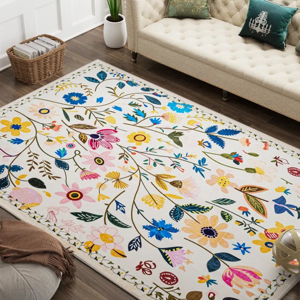 A floral-patterned rug with a variety of colorful flowers is placed on a wooden floor, adding a vibrant touch to a cozy room with a cream-colored sofa, decorative pillows, a potted plant, and a basket.