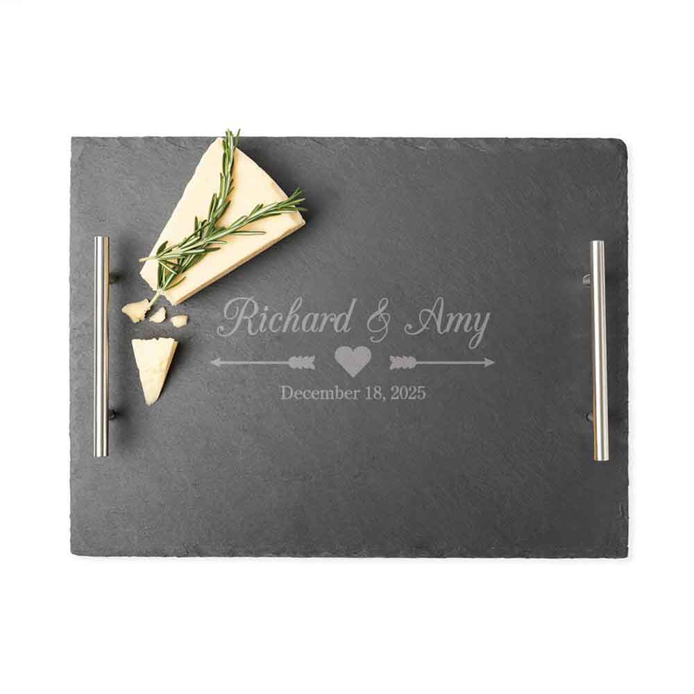A black slate tray with metal handles features the personalized engraving \“Richard & Amy\“ above a heart and arrow design, along with the date \“December 18, 2025.\“ It includes a piece of cheese garnished with rosemary.
