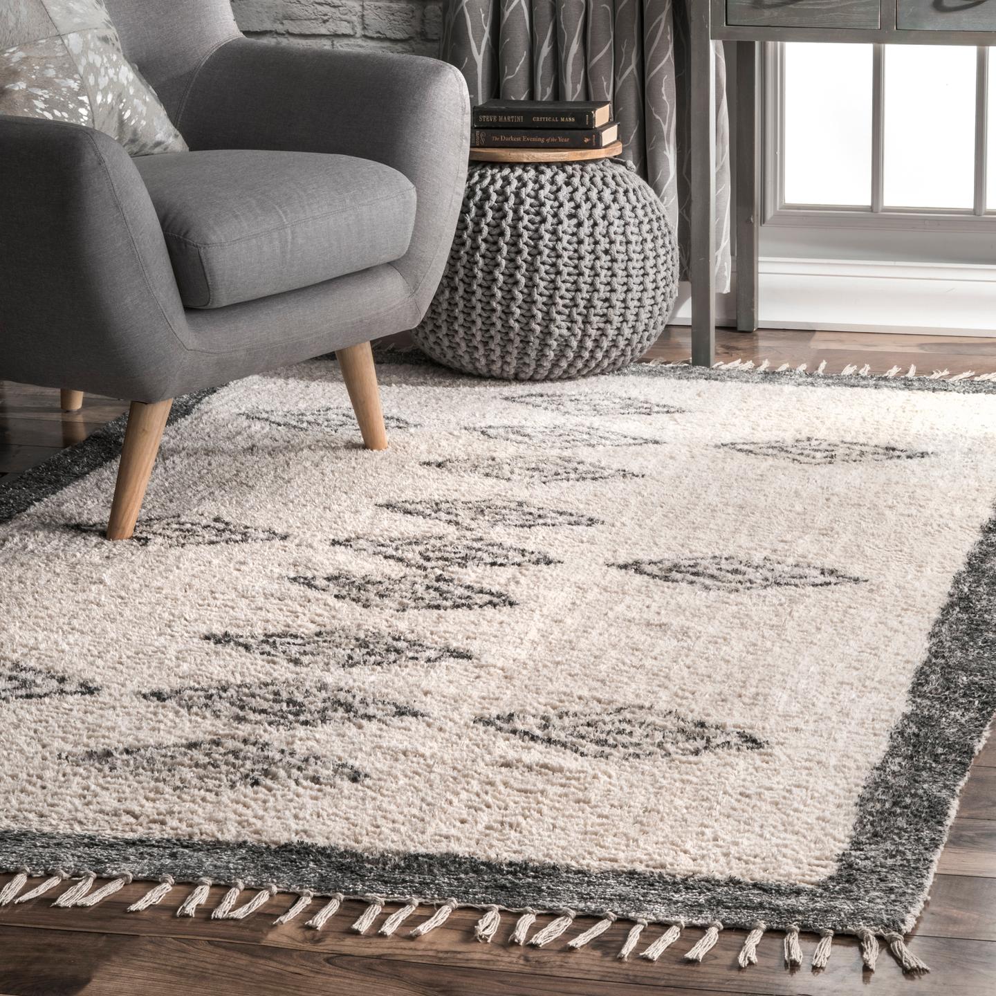 A gray upholstered chair with wooden legs sits next to a knitted pouf and a patterned rug with fringed edges on a hardwood floor.