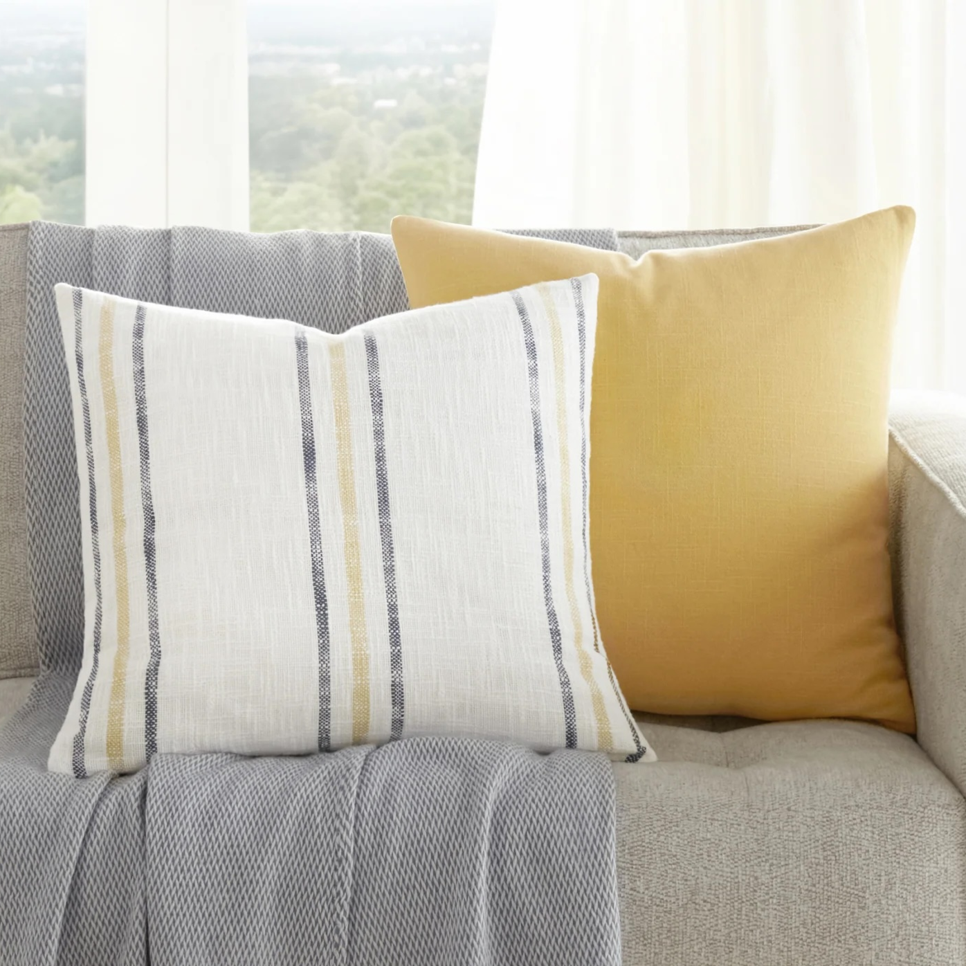 A beige sofa with a gray throw and two cushions, one with yellow stripes and the other solid yellow, is next to a small side table with a plant.