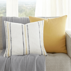 A beige sofa with a gray throw and two cushions, one with yellow stripes and the other solid yellow, is next to a small side table with a plant.