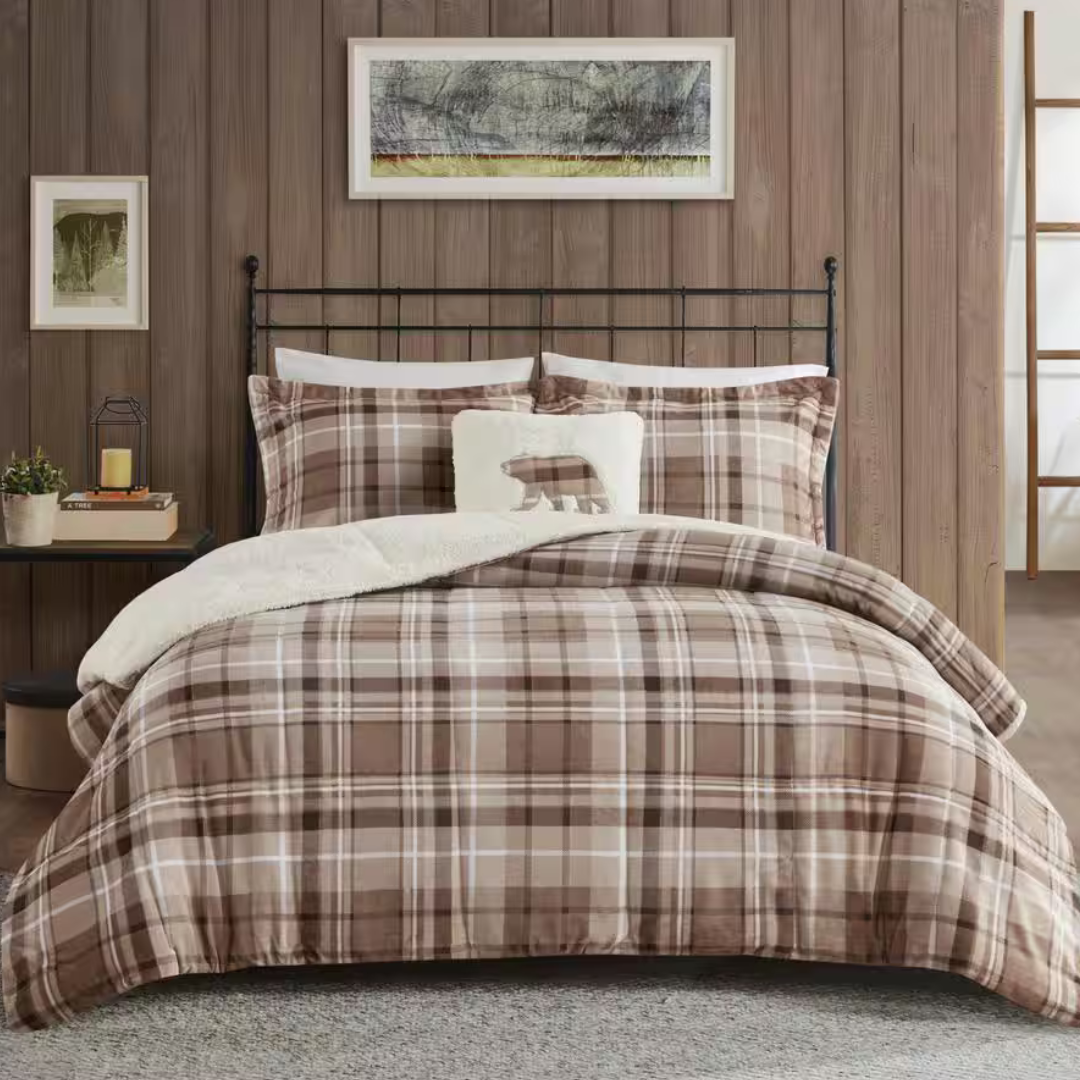 Plaid bedding set with brown tones, featuring a fluffy white pillow with a bear silhouette. The setting includes wooden panel walls, a metal bed frame, side table with a candle and plant, and framed wall art.
