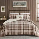 Plaid bedding set with brown tones, featuring a fluffy white pillow with a bear silhouette. The setting includes wooden panel walls, a metal bed frame, side table with a candle and plant, and framed wall art.
