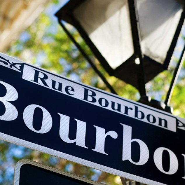 Street sign for Rue Bourbon, also known as Bourbon Street, in New Orleans. A classic black and white design featuring decorative elements with a traditional streetlamp in the background.