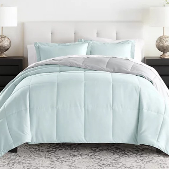 A light blue reversible comforter set features a stitched grid pattern, accompanied by matching pillow shams. It sits atop a bed with a cushioned white headboard, flanked by two black nightstands and lamps with textured bases.
