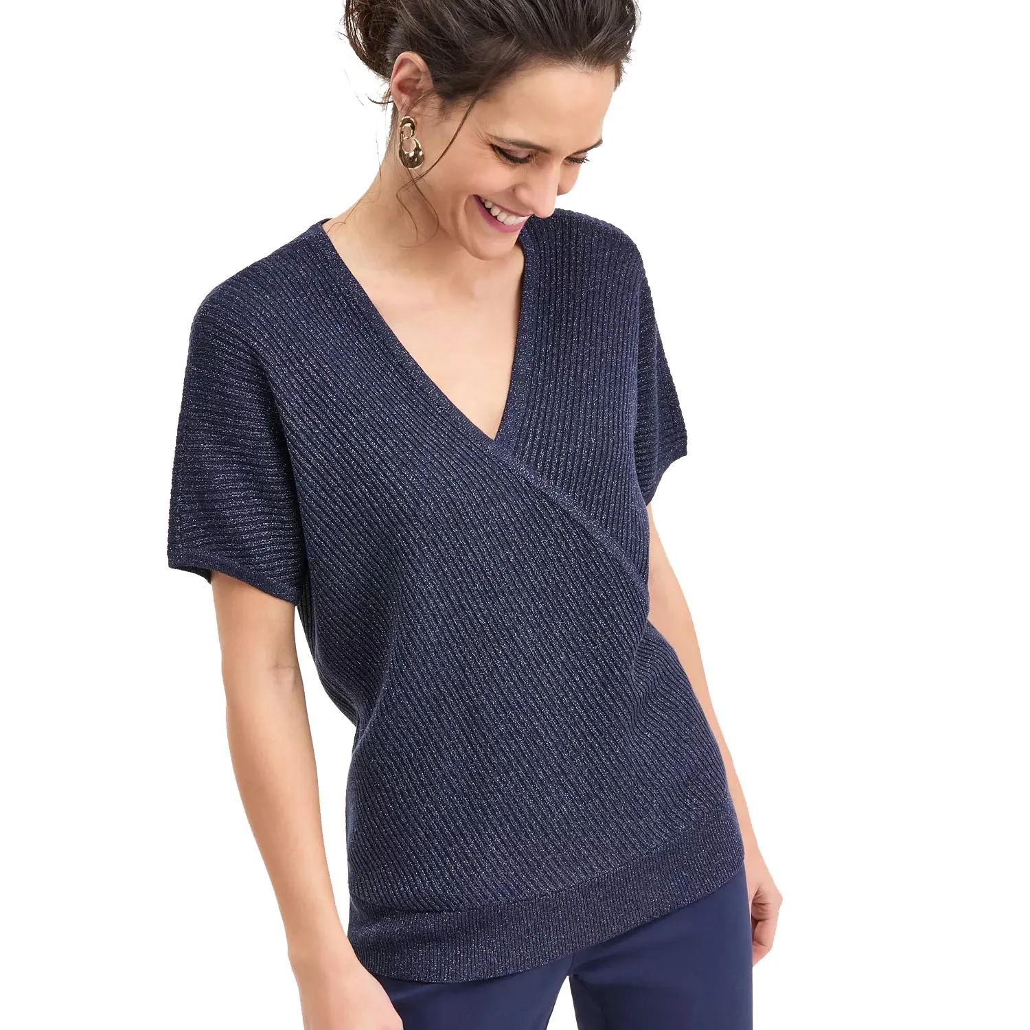 A woman wearing a short-sleeve, deep V-neck, textured navy wrap-style top paired with navy pants. She has hoop earrings and her hair styled up.