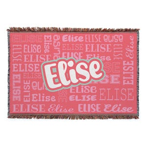 A woven blanket with fringed edges features the name \“Elise\“ in various fonts and sizes against a pink background.
