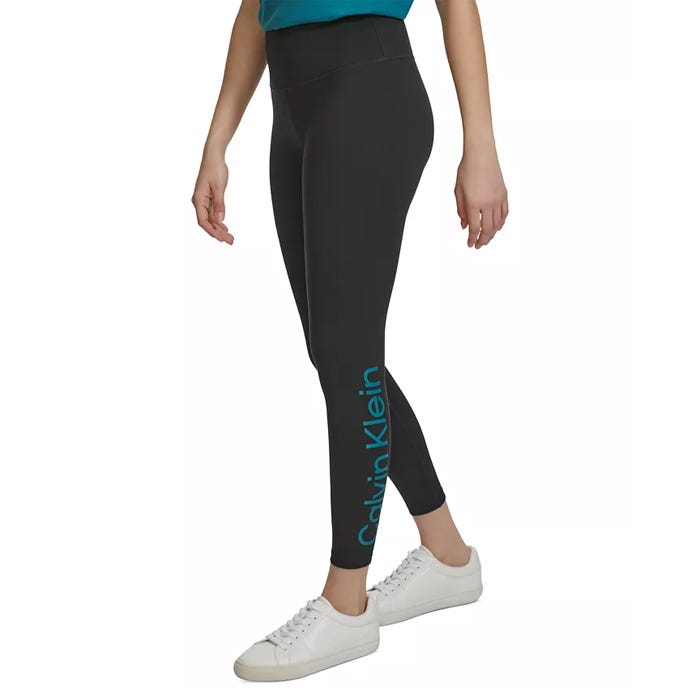 High-rise 7/8 leggings in a dark shade, featuring the Calvin Klein logo on the lower left leg and paired with white sneakers.