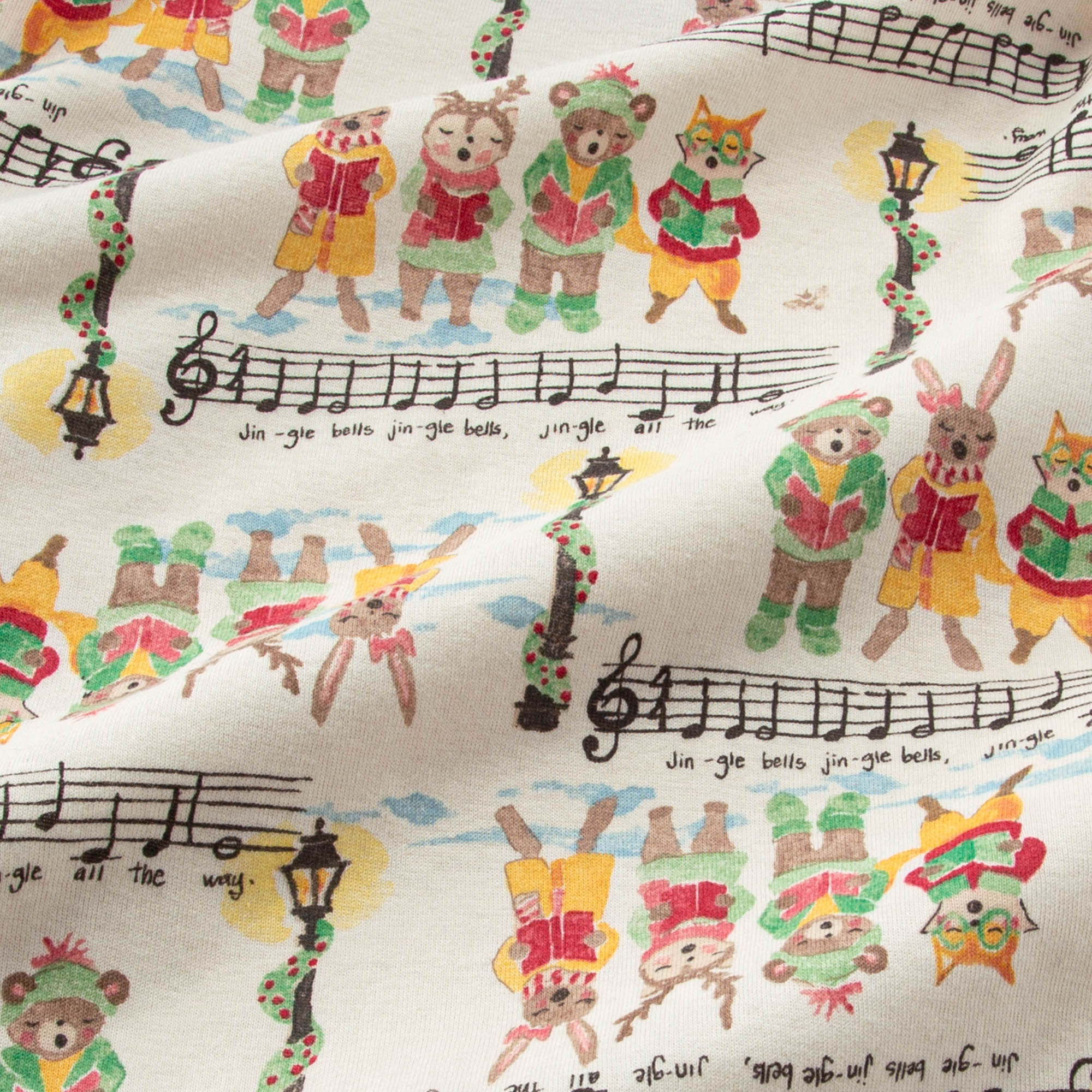 Fabric with a whimsical holiday print features animals dressed in festive outfits reading from songbooks. It includes musical notes and lyrics from \“Jingle Bells\“ along with decorated street lamps.