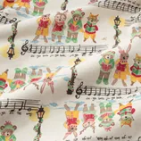 Fabric with a whimsical holiday print features animals dressed in festive outfits reading from songbooks. It includes musical notes and lyrics from \“Jingle Bells\“ along with decorated street lamps.